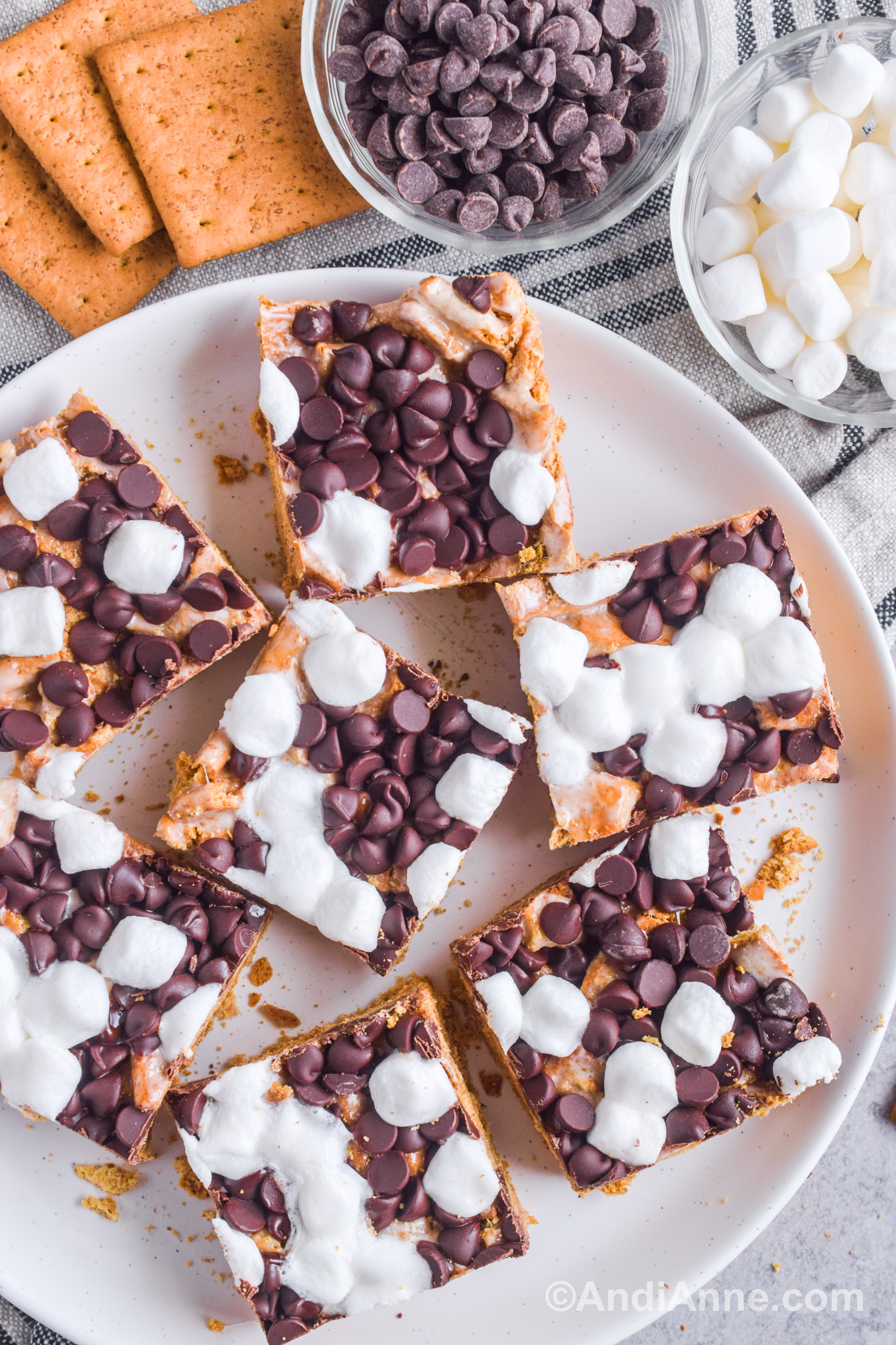 A plate of square s'more bars with bowl of chocolate chips, bowl of mini marshmallows and graham crackers surrounding them.