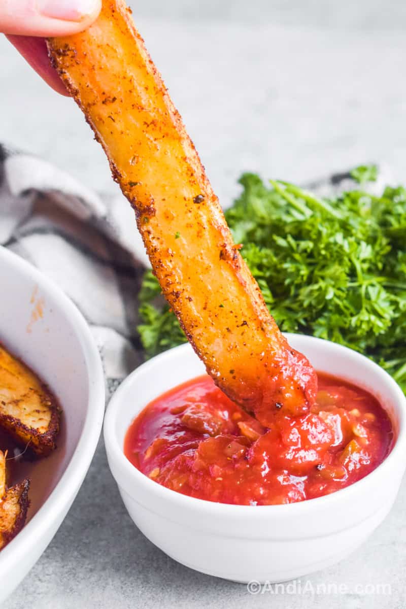 Fingers dipping a baked potato wedge in a white bowl of salsa. Fresh green parsley is in the background.