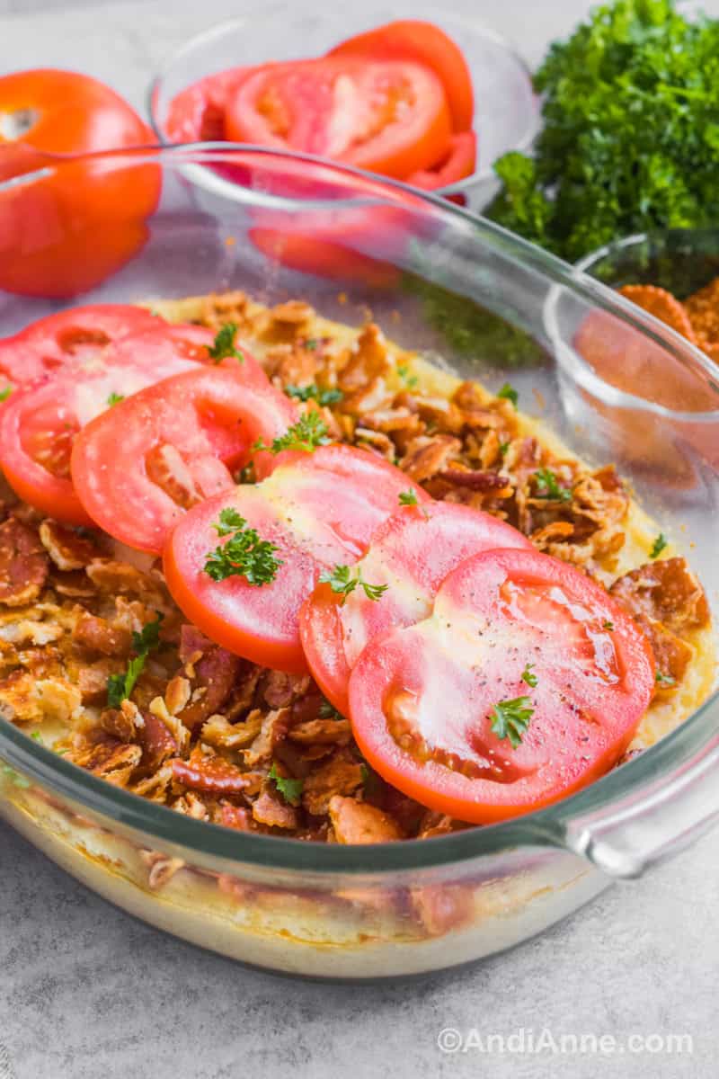A glass dish of mashed turnips, crushed crackers and tomato slices. Behind is bowl of tomato slices, crackers and parsley.