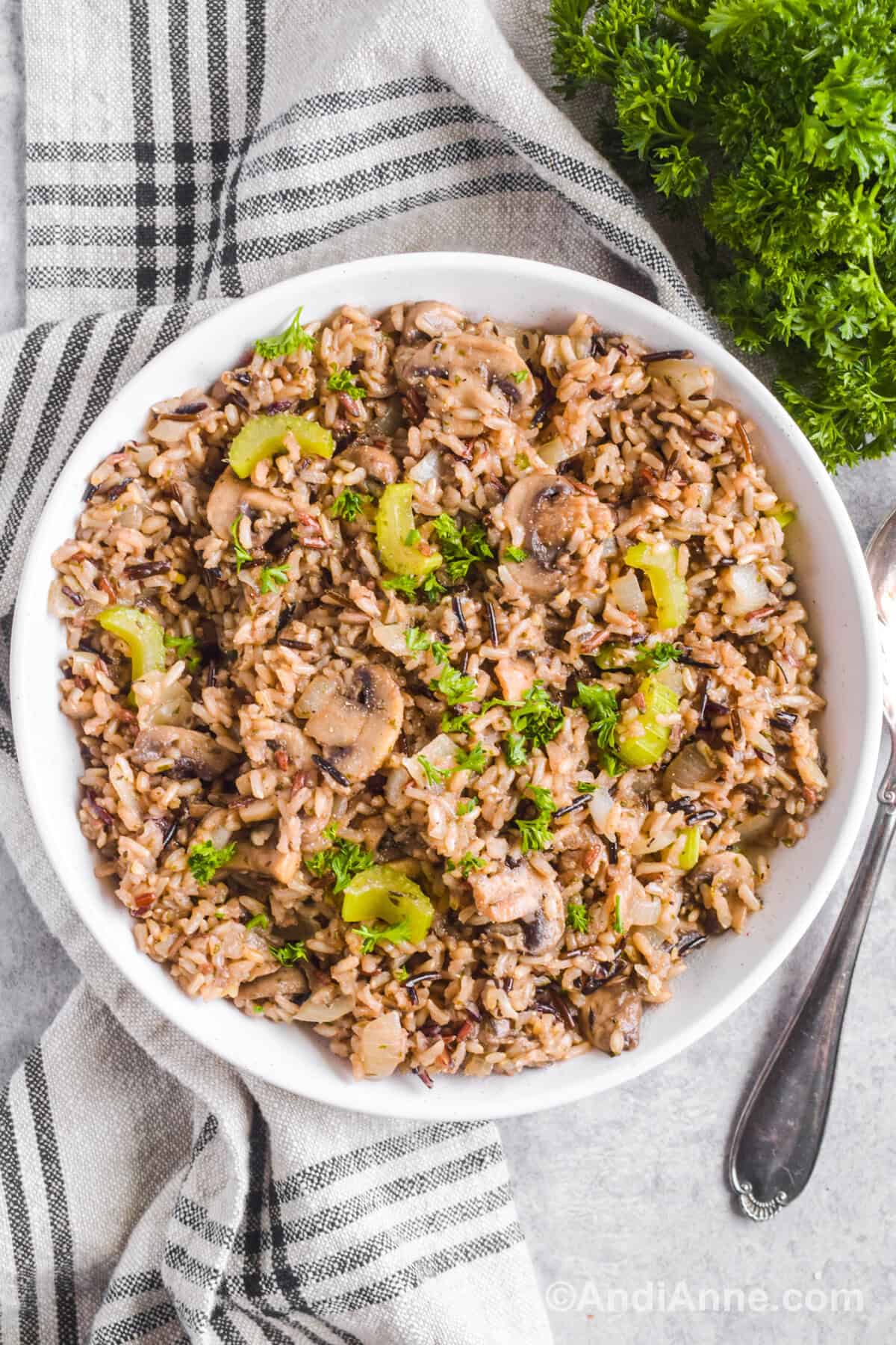Looking down at a white bowl of wild rice stuffing with parsley and kitchen towel surrounding dish.
