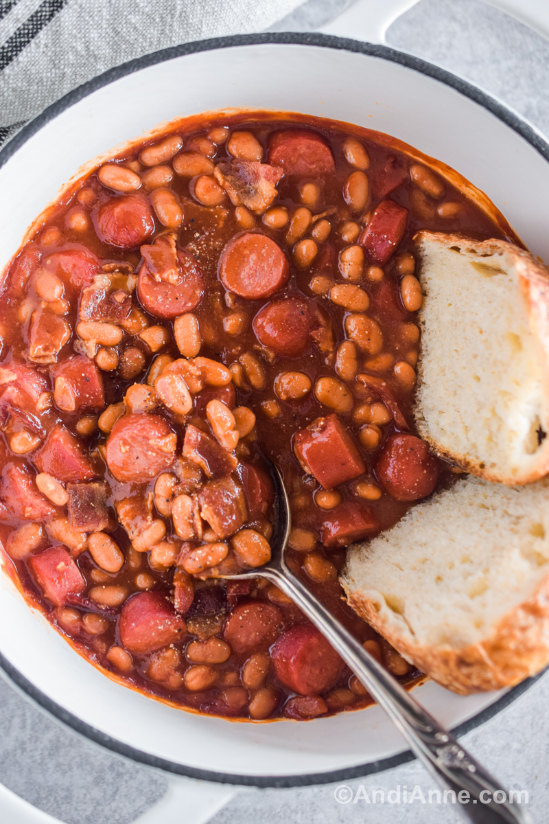 Close up of a white pot with beans and franks recipe. A big spoon and slices of bread in the pot. 