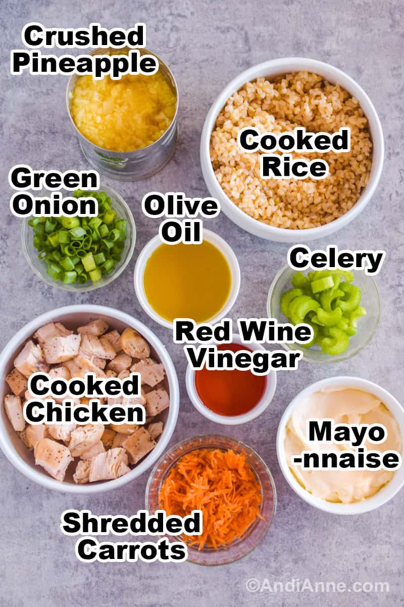 Recipe ingredients on a counter including white bowl of cooked rice, chopped cooked chicken, bowls of green onion, olive oil, red wine vinegar, celery, mayonnaise, and shredded carrots.