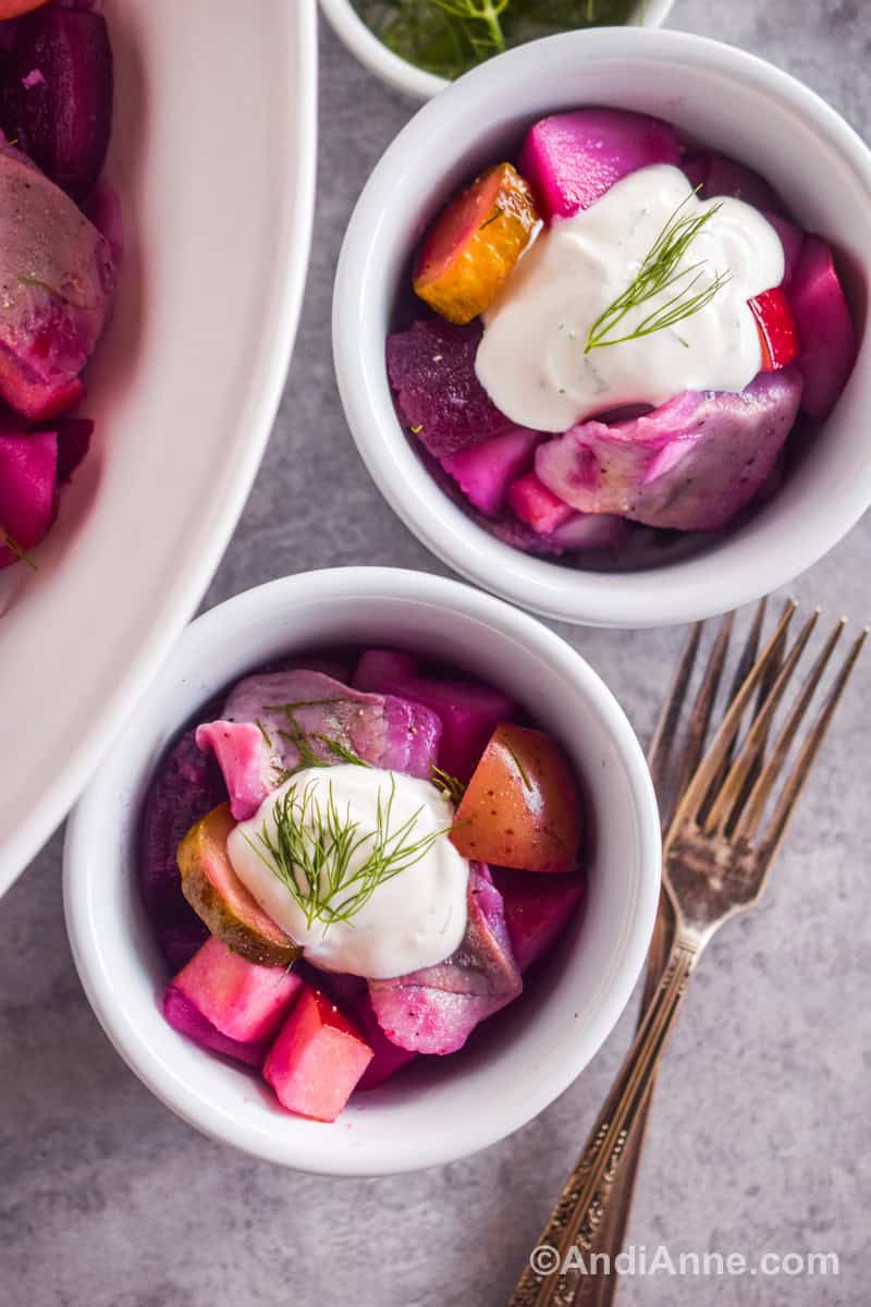 Two small bowls with herring salad with beets and pickles. A dollop of dill cream sauce on top and a small sprig of fresh dill. Forks and a large bowl of salad surround the bowls.