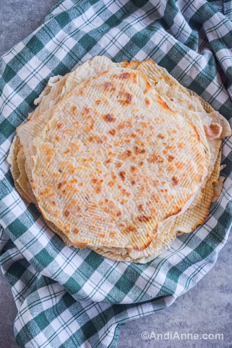 A pile of cooked lefse on a plate with a green and white kitchen towel underneath.