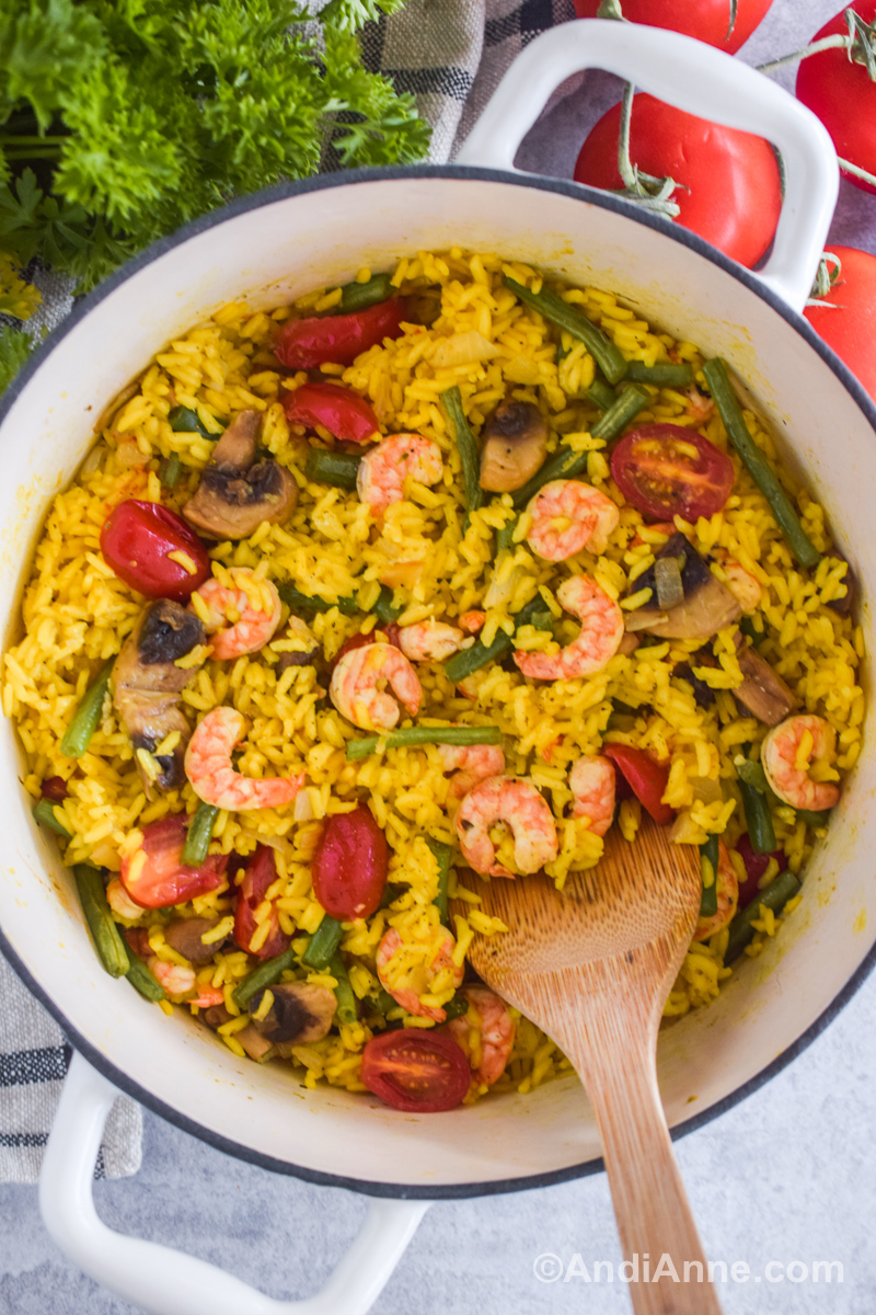 A large white pot with yellow rice, shrimp, green beans and mushrooms. A wood spatula sits inside, and parsley and tomatoes surround the pot.