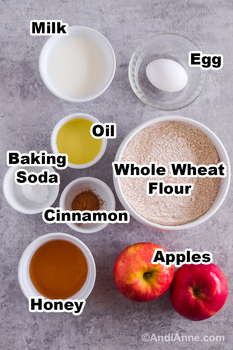 Recipe ingredients on a counter including bowls of milk, flour, oil, cinnamon, honey, baking soda and two apples.