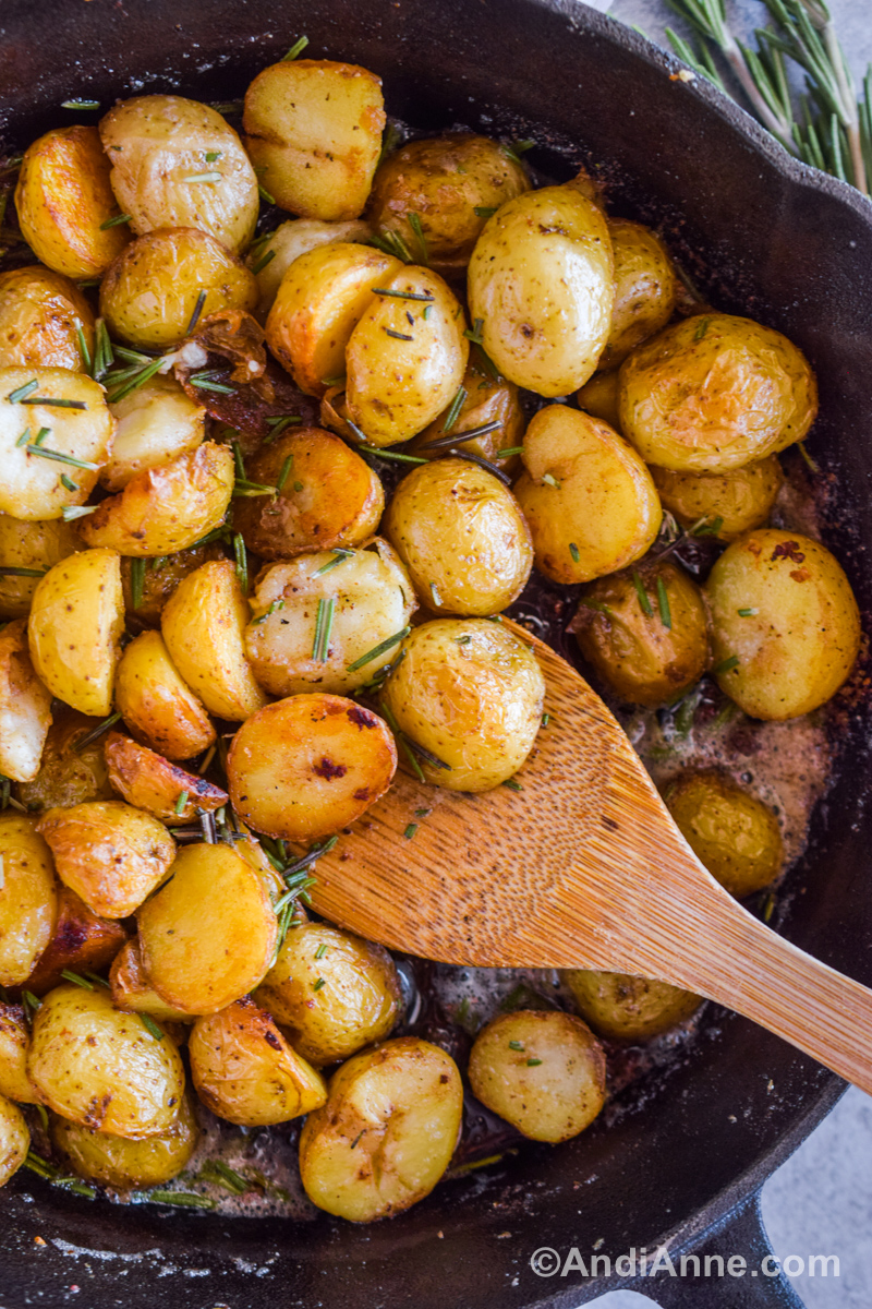 Crispy baby potatoes in a skillet with chopped rosemary and a wood spoon.