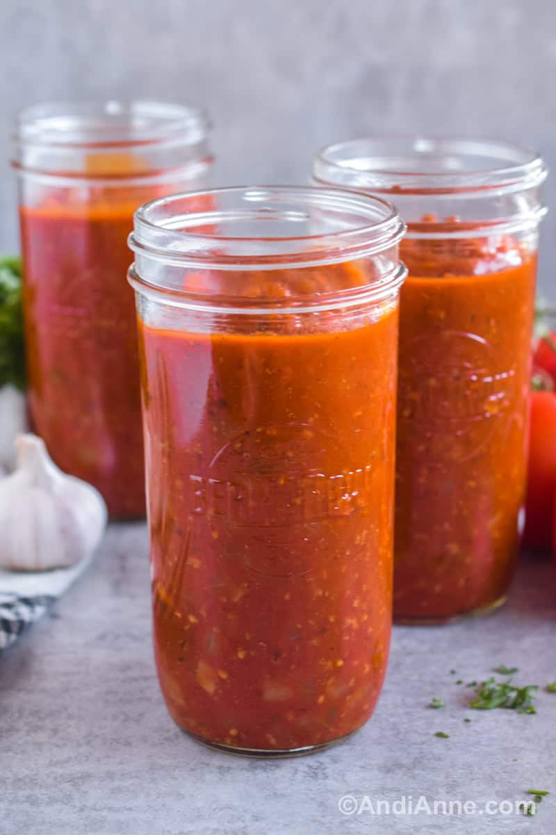 Three mason jars of spaghetti sauce on a counter together with garlic and tomatoes in the background.
