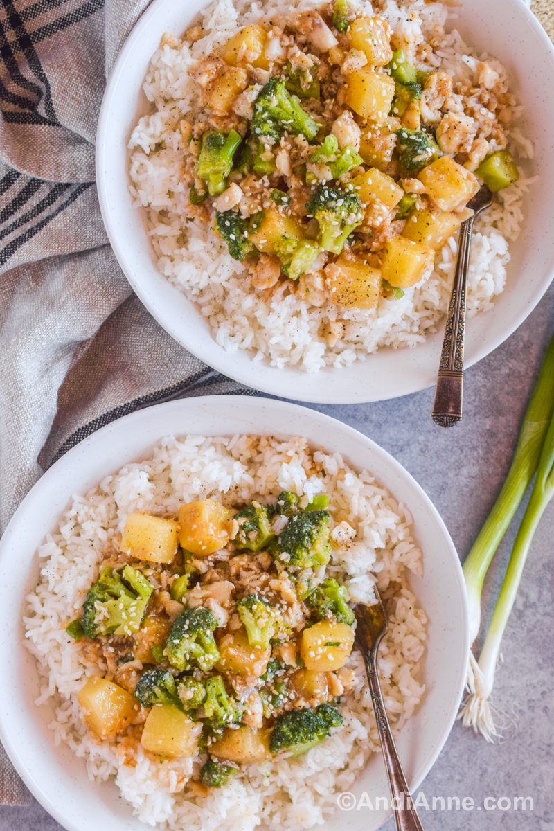 Two white bowls with rice and topped with pineapple chunks, broccoli and fish in a brown sauce and topped with sesame seeds. 