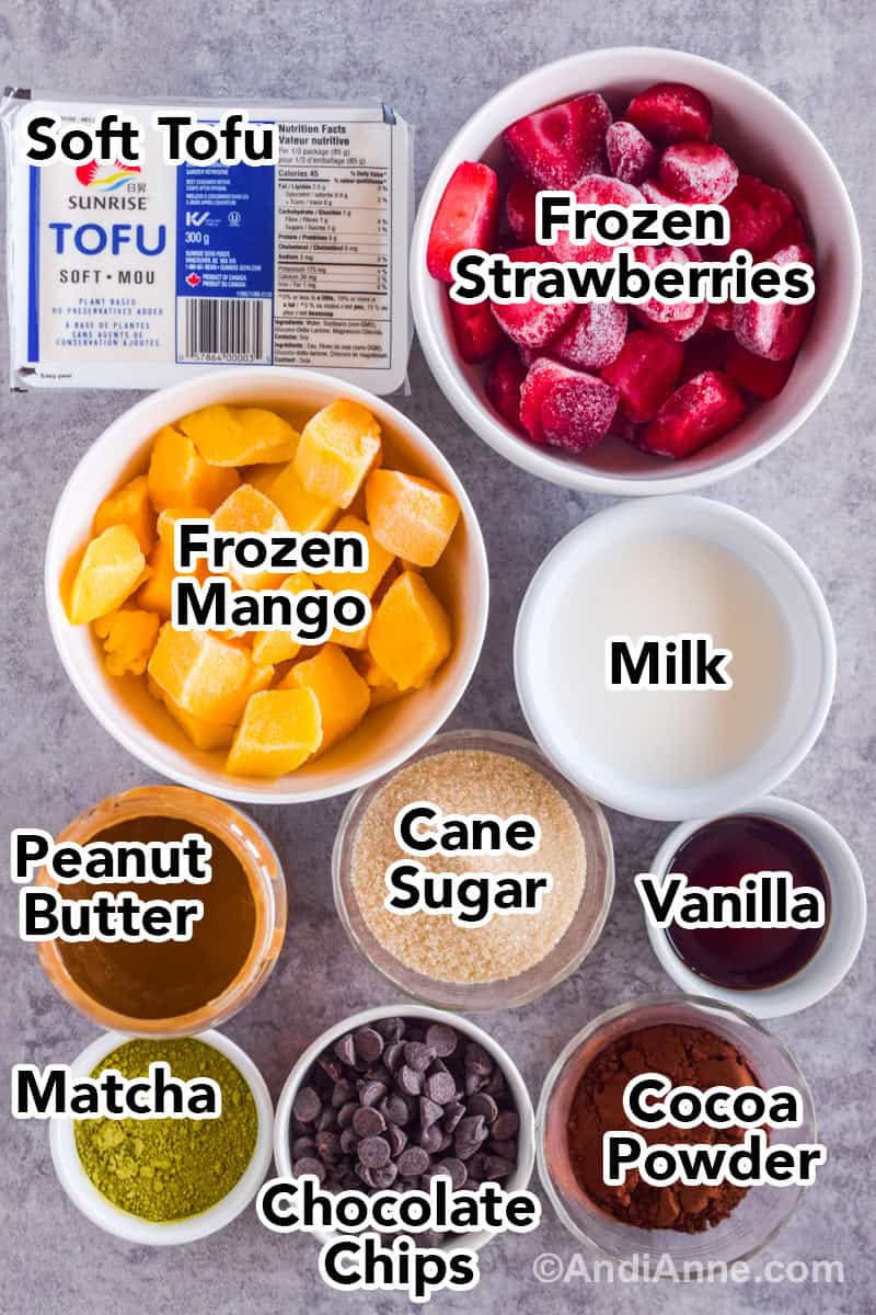 Looking down at recipe ingredients on a table including bowls of frozen mango, strawberries, bowls of milk, peanut butter, sugar, vanilla, matcha powder, chocolate chips and cocoa powder.