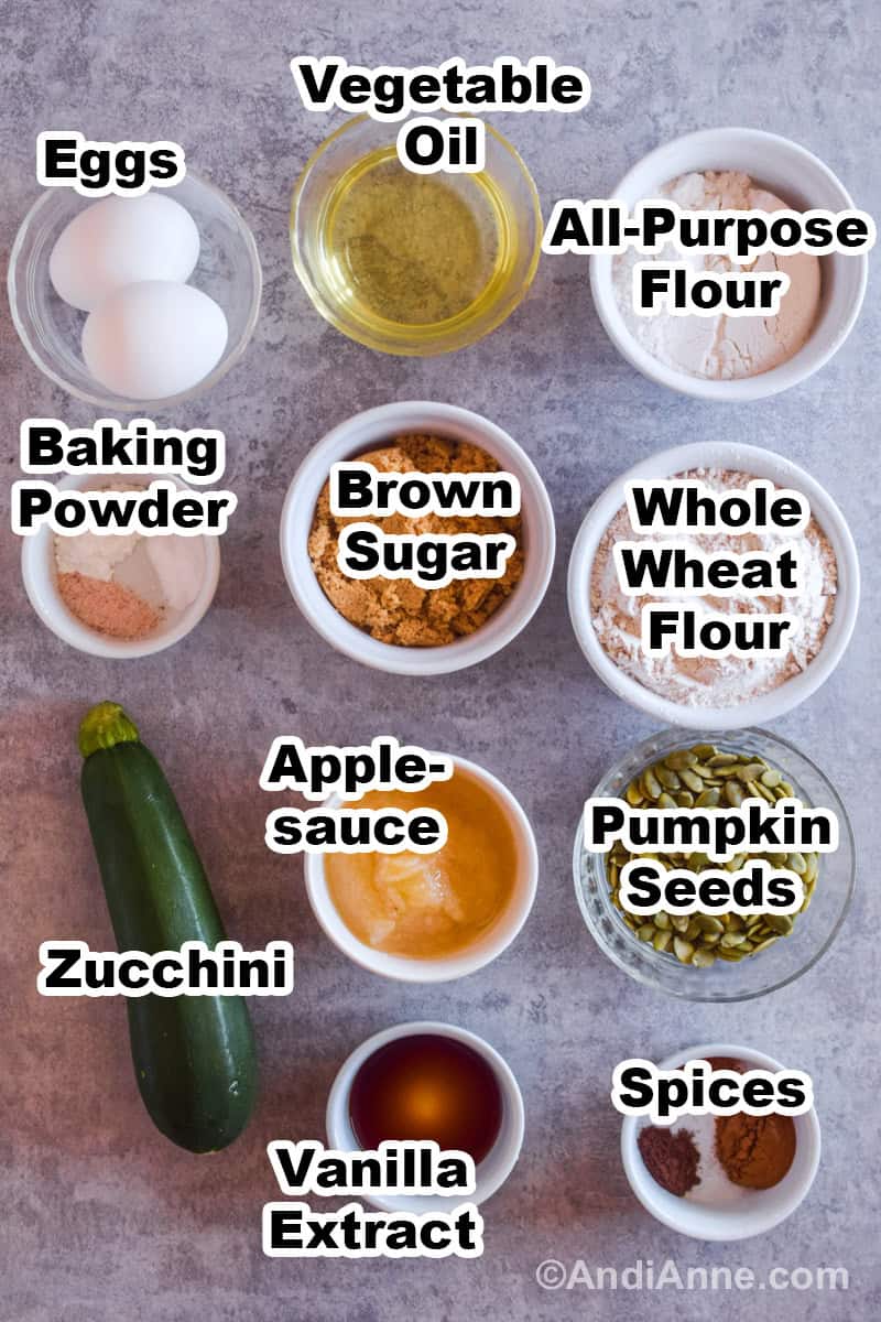 Ingredients to make recipe on a counter including bowls of flour, sugar, seeds, applesauce, spices, vanilla and a zucchini.