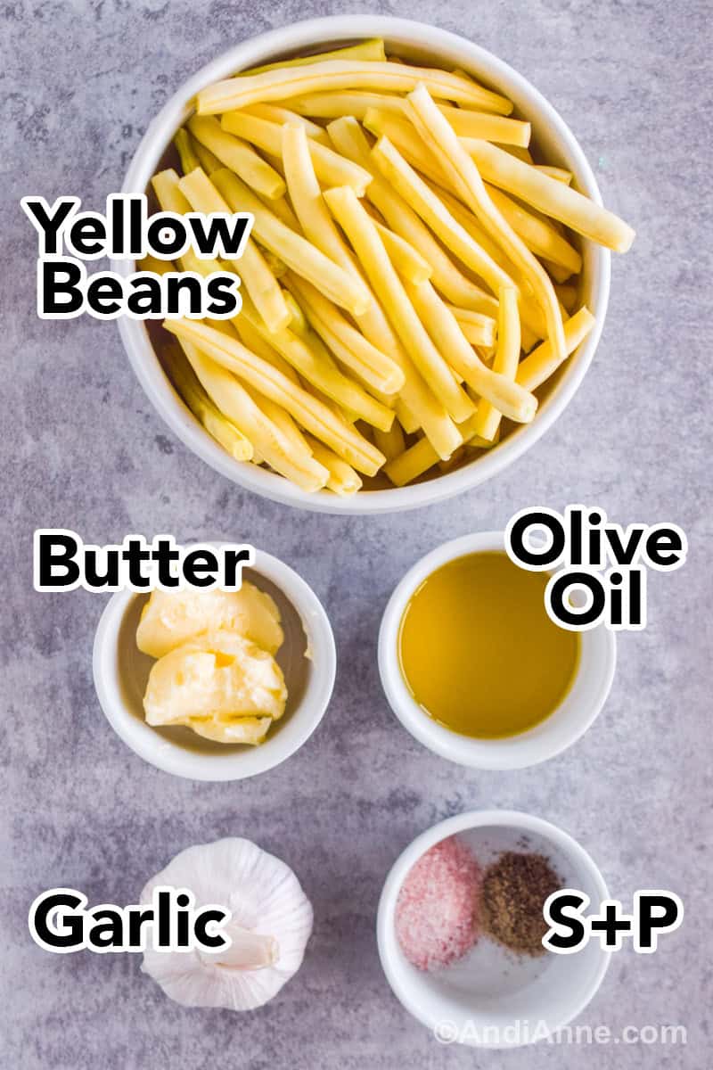 Ingredients for the recipe including a bowl of yellow beans with trimmed edges, bowls of butter, olive oil, salt and pepper, and a head of garlic.