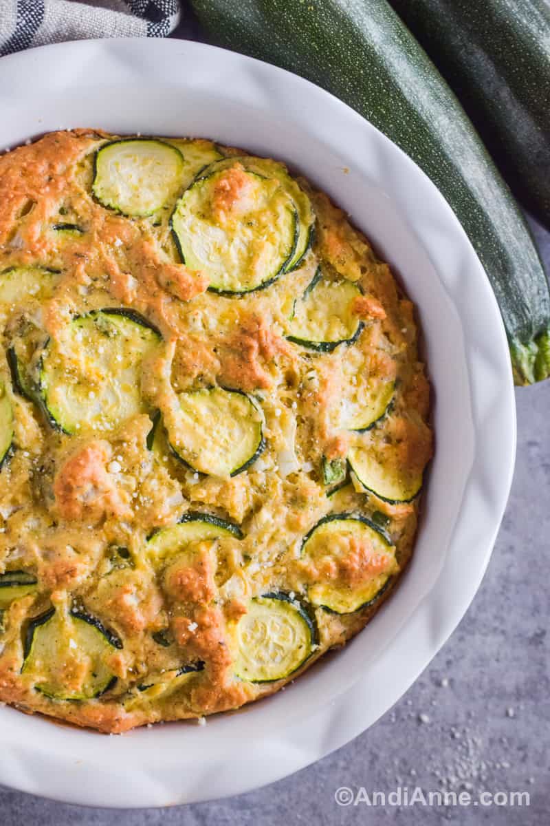 Close up of sliced zucchinis in a baked dough inside a white pie plate with zucchinis beside it.