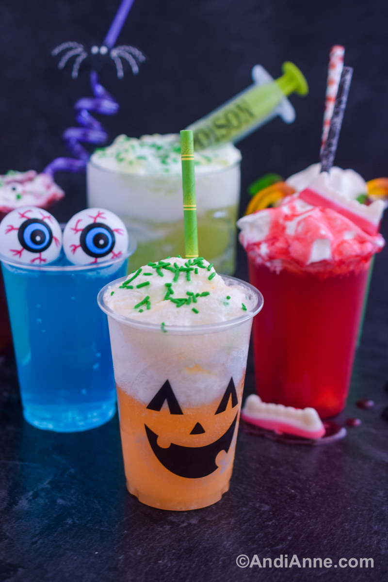 Four different halloween drinks in plastic cups in different colors with various toppings including sprinkles, whipped cream, plastic eyeballs and strawberry syrup.