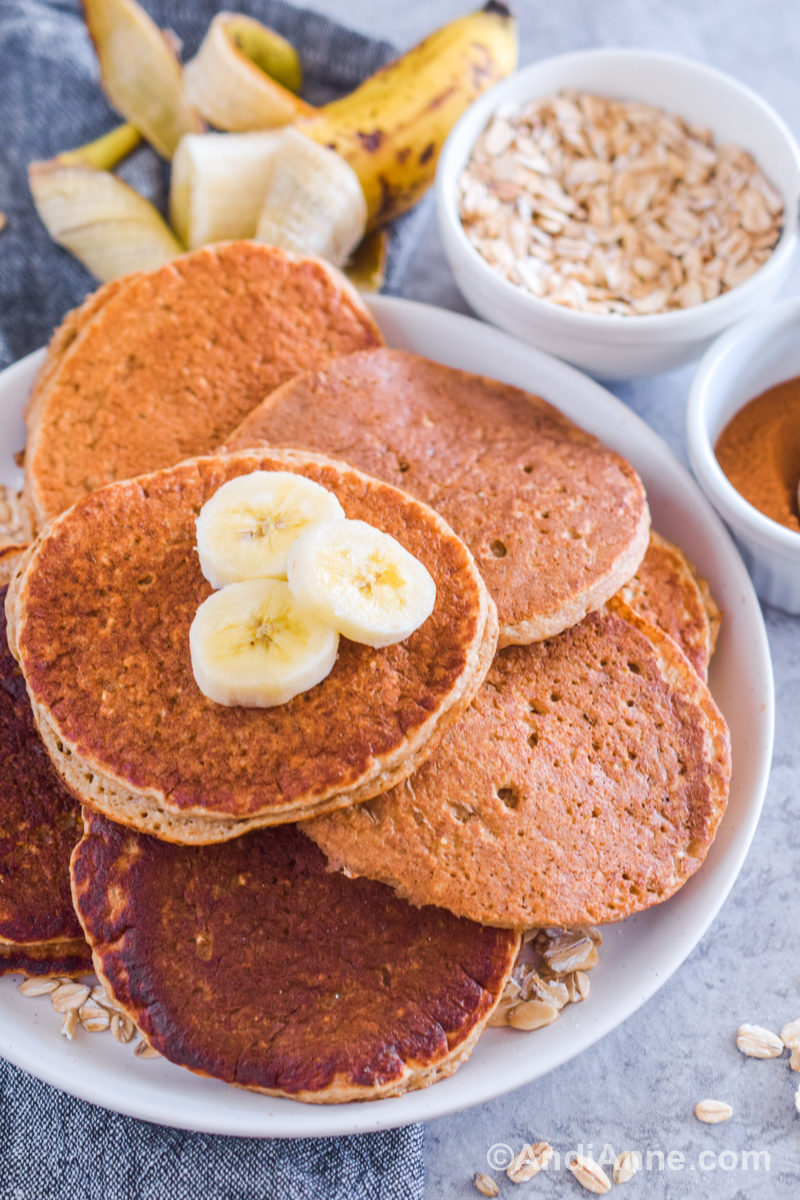 A pile of oatmeal pancakes on a plate with slices of banana. Raw banana and bowls of oats and cinnamon in the background. 