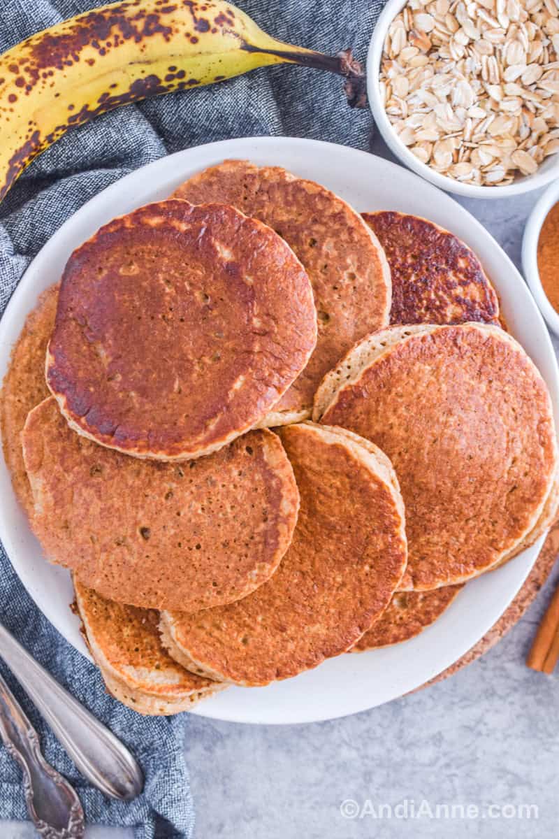 Cooked banana oatmeal pancakes on a white plate with a ripe banana, bowl of rolled oats and cinnamon in the background.