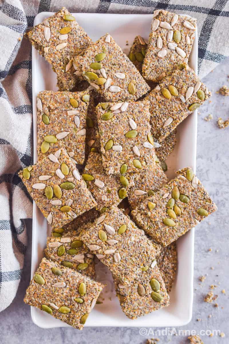 Seed crackers piled on top of a white rectangle plate with a kitchen towel behind it.