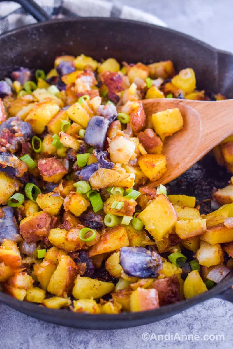 close up of chopped potatoes, green onions and spices in hash browns in a skillet with a wood spoon.