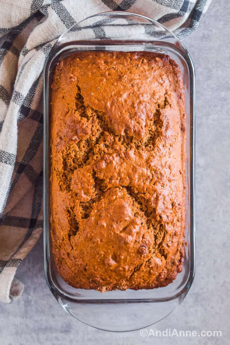 Close up of baked brown bread in a glass loaf pan with a plaid kitchen towel beside it.