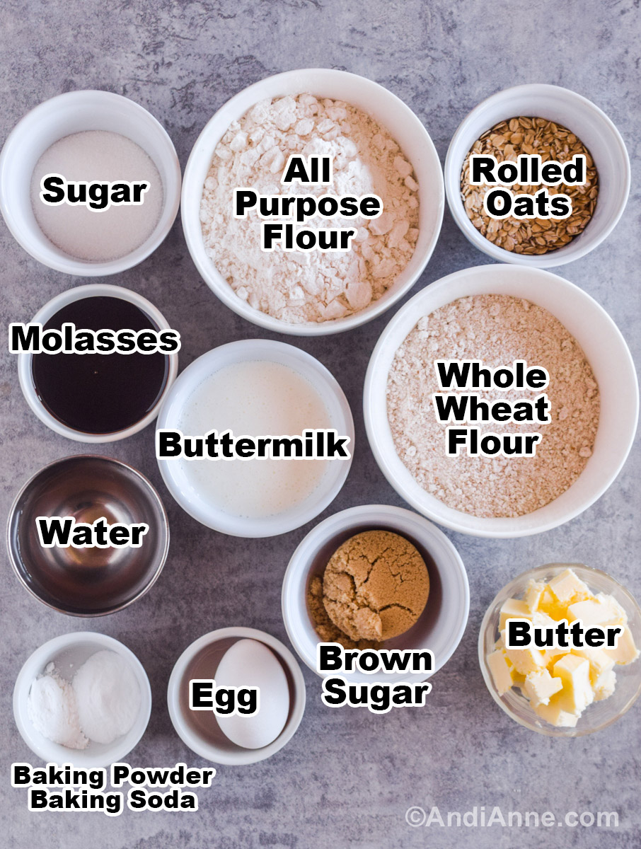 Recipe ingredients on a counter in bowls including all purpose flour, whole wheat flour, buttermilk, molasses, sugar, rolled oats, water, brown sugar, butter and egg.