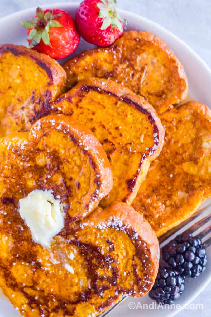 Close up of three pieces of french toast on a white plate with blackberries and blueberries. Drizzled with maple syrup and sprinkled with powdered icing sugar.