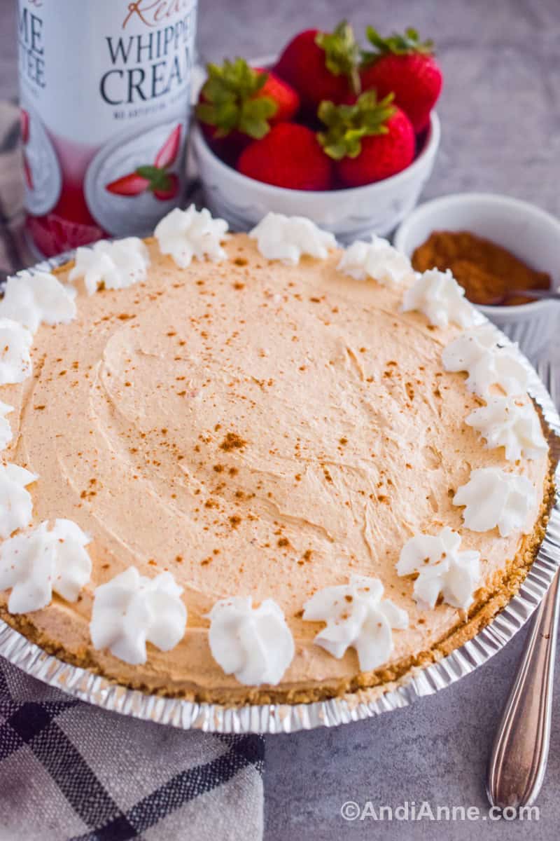 Looking on an angle at the pumpkin marshmallow pie with strawberries, canned whipped cream and a bowl of cinnamon in the background.