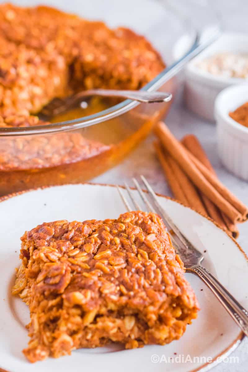 Close up of one slice of baked oats on a small plate with a fork. Cinnamon sticks, bowls and the serving dish with rest of recipe are behind it.