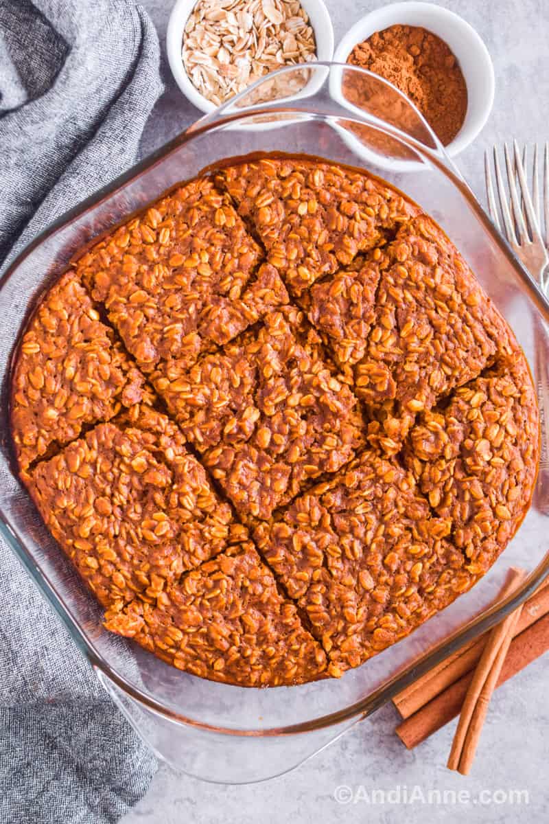 Square glass baking dish with baked pumpkin spiced oats inside. Cinnamon sticks, forks and small bowls of oats and cinnamon surround the dish.