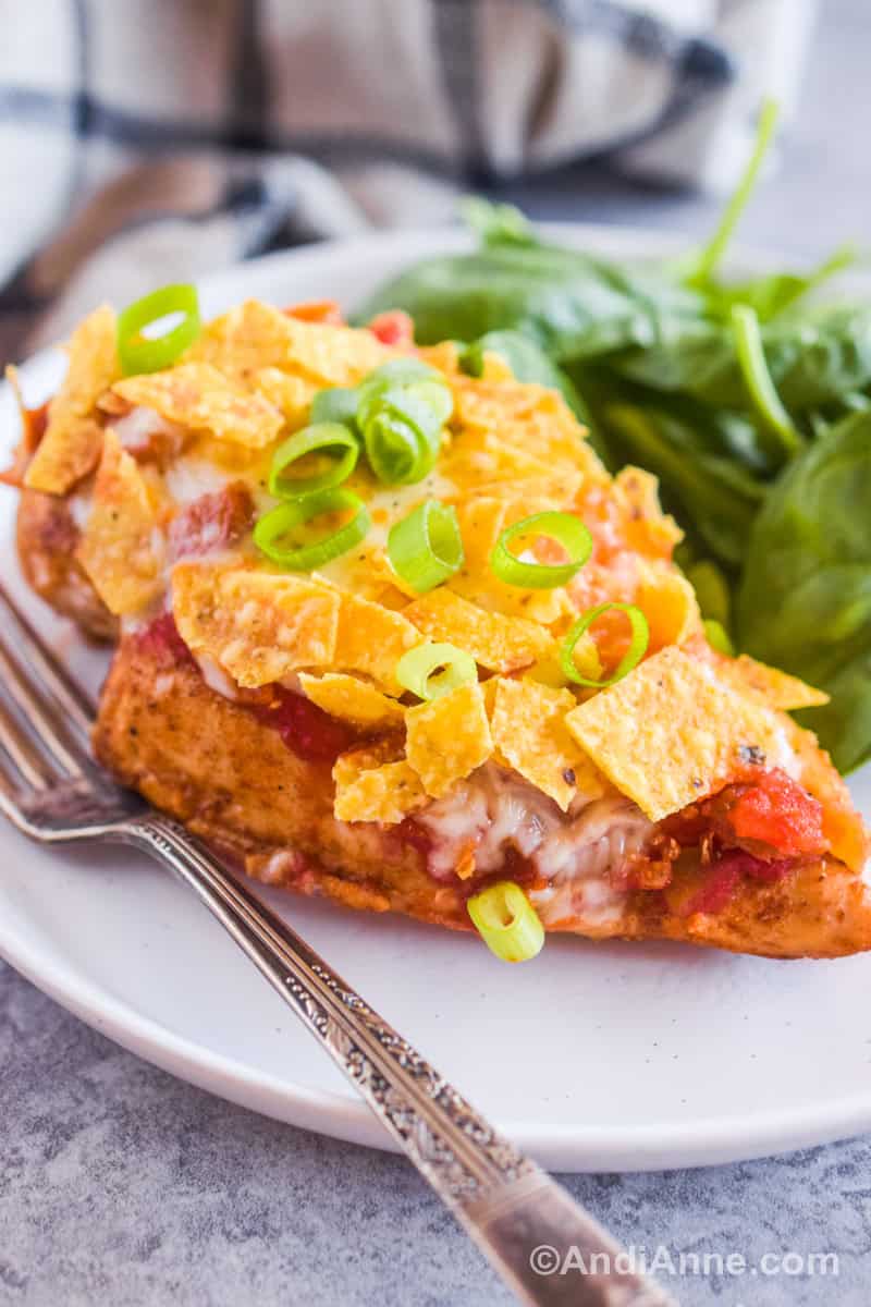 Close up of one chicken breast covered in salsa, cheese, crushed tortilla chips and chopped green onion with a pile of spinach in the background.