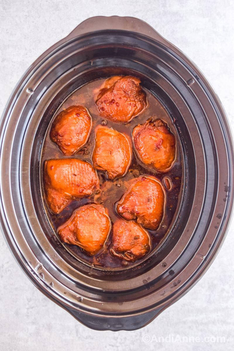 Looking down at a slow cooker with sauce and brown colored chicken thighs inside.