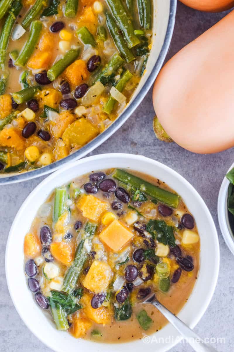 A white bowl with vegetable soup and as spoon. A large pot with more soup and a butternut squash beside it.