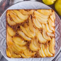 Upside down pear gingerbread cake on a white plate with pears and a bowl of cinnamon beside it.