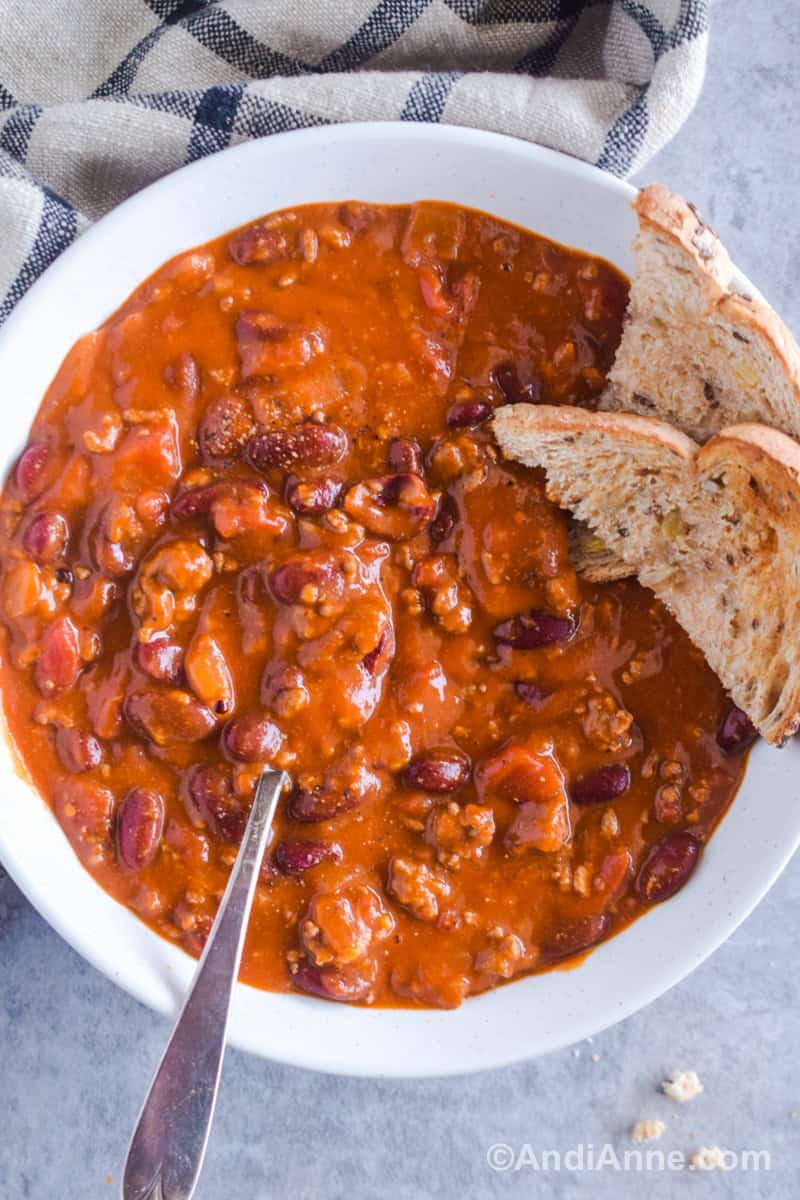 A white bowl with chili and a spoon. Slices of toast are on the side of the bowl.