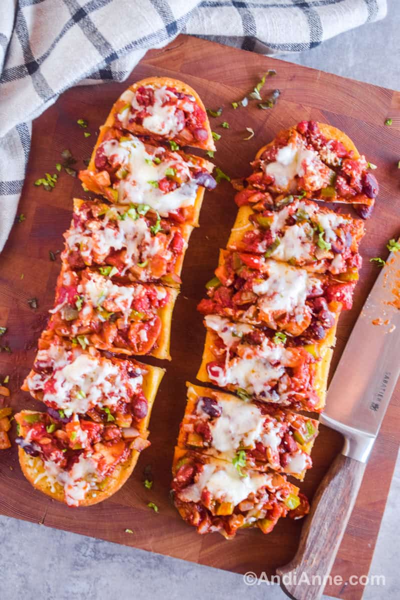 Sliced garlic bread topped with chili mixture and melted cheese on a cutting board with a knife beside.
