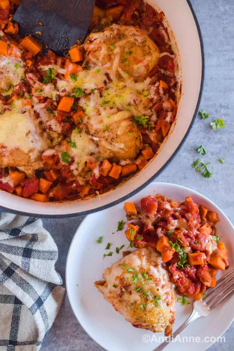 A plate with chicken thigh and cooked sweet potatoes and tomatoes. A pot with cooked ingredients from recipe beside the plate.