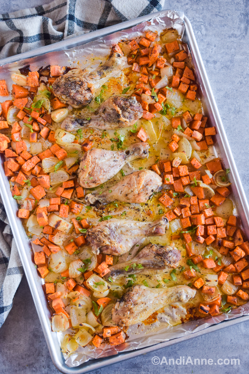 Looking down at cooked chicken legs, chopped sweet potato and onion on a baking sheet.